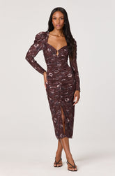 A woman is wearing a long, fitted dress with a dark brown base color and a floral pattern in shades of light purple and gray. The dress features long sleeves with puffed shoulders, a sweetheart neckline with a small keyhole cutout in the center, and gathers along the torso for a ruched effect. It also has a slit at the front of the skirt, reaching to mid-calf. She is wearing strappy black high-heeled sandals.